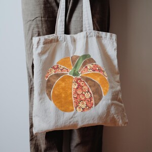 May include: A beige cotton tote bag featuring a large, central pumpkin applique. The pumpkin is constructed from alternating fabric segments in solid mustard yellow, dark brown, and a floral print pattern, topped with a green fabric stem.