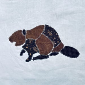 May include: A white fabric square featuring a central applique of a beaver. The beaver is constructed from various brown and dark brown patterned fabric patches, joined together with visible white zigzag stitching along the edges.