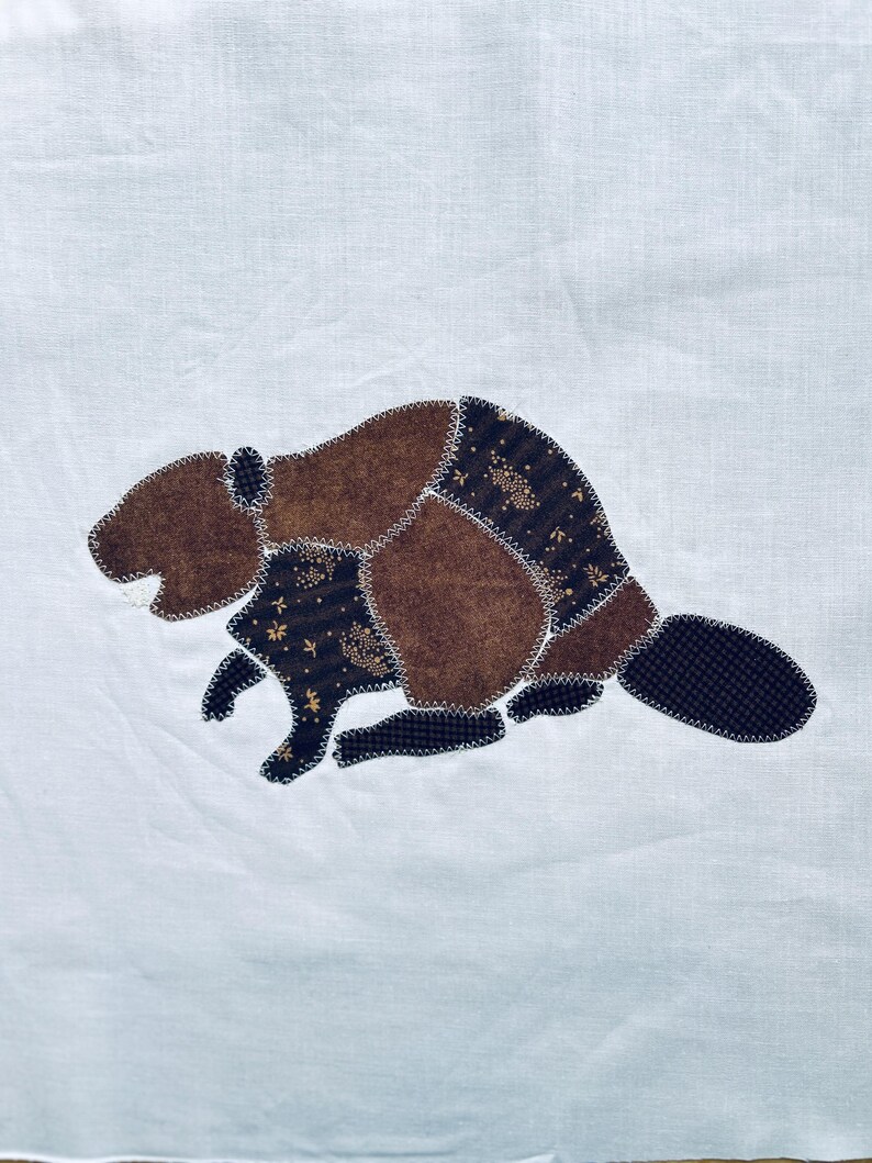 May include: A white fabric square featuring a central applique of a beaver. The beaver is constructed from various brown and dark brown patterned fabric patches, joined together with visible white zigzag stitching along the edges.