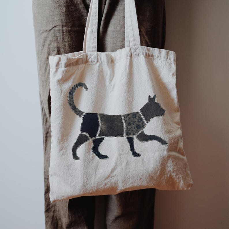 May include: A cream-colored canvas tote bag featuring a large, stylized cat graphic on the front. The cat is composed of several dark, patterned fabric segments stitched together with visible white thread, creating a patchwork effect.