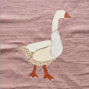 May include: A fabric appliqué of a goose on a dusty rose textured background. The goose is constructed from various cream and beige patterned fabric pieces, including a tan gingham tail and wing section, with a solid orange beak and webbed feet.