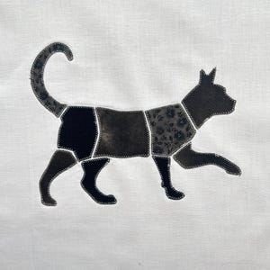 May include: A white fabric panel featuring a cat silhouette appliqu&eacute;d in sections. The cat is composed of various black and dark gray fabric pieces with different textures and floral patterns, all joined together with visible white zigzag stitching.