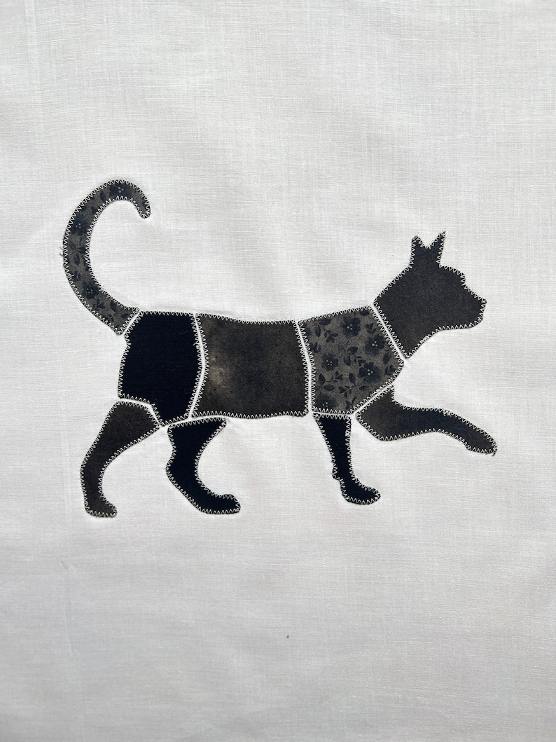 May include: A white fabric panel featuring a cat silhouette appliqu&eacute;d in sections. The cat is composed of various black and dark gray fabric pieces with different textures and floral patterns, all joined together with visible white zigzag stitching.