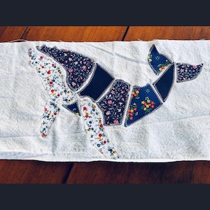 May include: A rectangular piece of white fabric featuring a whale applique constructed from various fabric patches. The whale is composed of dark blue, white, and floral patterned segments joined together with visible white zigzag stitching.