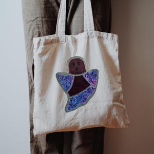 Op de afbeelding: Een cr&egrave;mekleurige canvas draagtas met een centrale geestvormige applicatie. De geest heeft een donker kastanjebruine kop en romp, met vleugels van paars en blauw patroonstof omlijnd door een glinsterende zilveren rand.