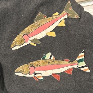 May include: A dark gray fleece fabric featuring two appliqu&eacute;d fish designs. The top fish has olive green, pink, and yellow patterned fabric sections. The bottom fish features similar colors with additional striped patterns on the fins and tail.