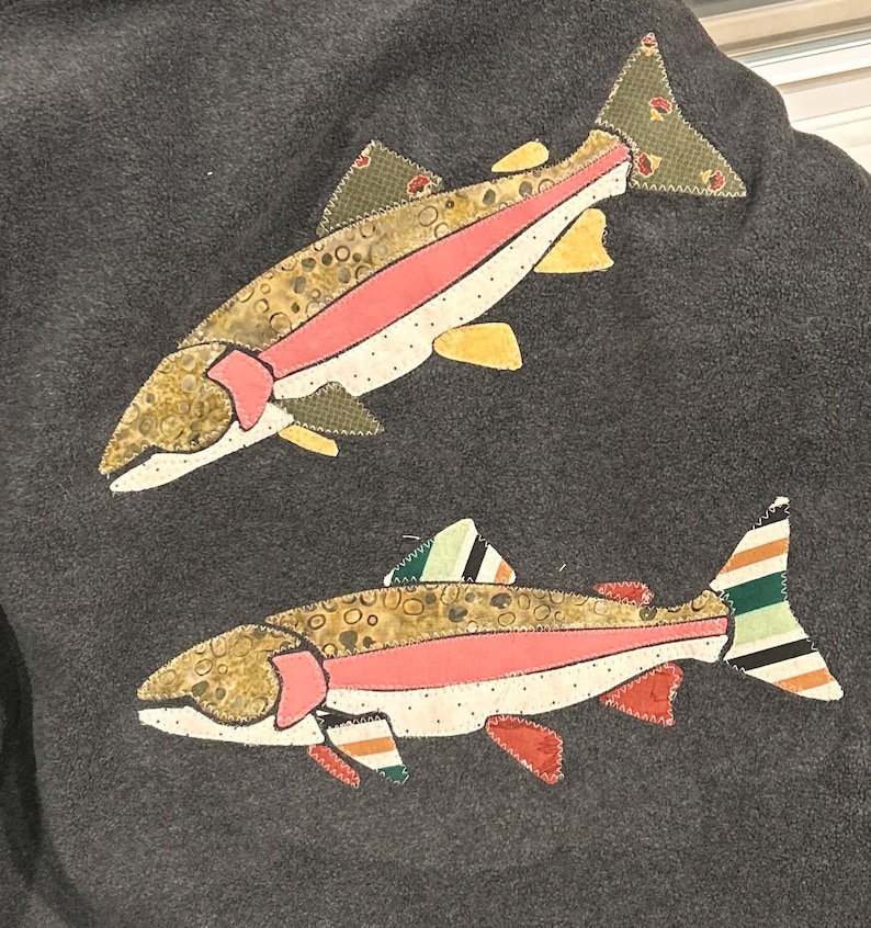 May include: A dark gray fleece fabric featuring two appliqu&eacute;d fish designs. The top fish has olive green, pink, and yellow patterned fabric sections. The bottom fish features similar colors with additional striped patterns on the fins and tail.