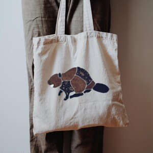 May include: A cream-colored canvas tote bag featuring a central applique of a beaver. The beaver is constructed from various fabric patches in shades of brown and dark blue with small star patterns, joined together by visible white stitching.