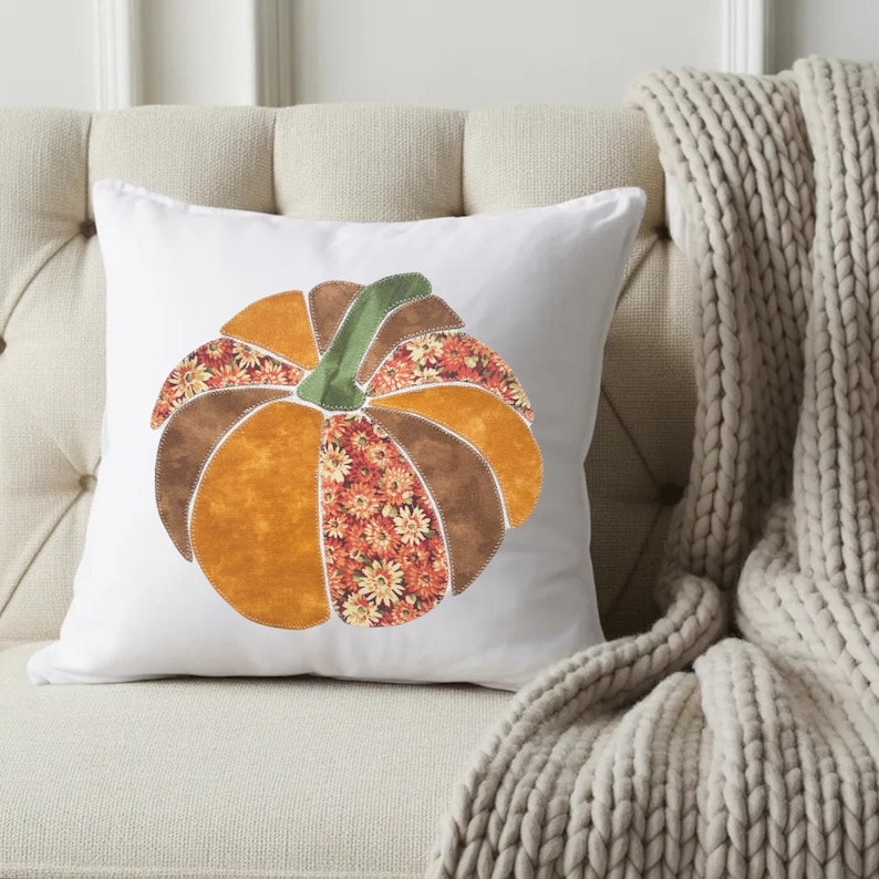May include: A square white throw pillow featuring a central pumpkin applique. The pumpkin is constructed from alternating fabric segments in solid orange, solid brown, and a floral print pattern of orange and yellow flowers with a green stem.