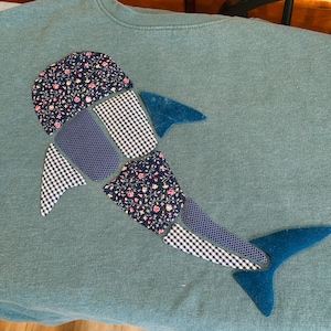 May include: A teal sweatshirt with a patchwork shark design. The shark is made from fabrics with floral, gingham, and chevron patterns in navy, white, and teal. The fins and tail are solid teal.