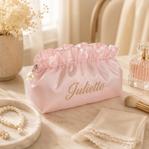 Peut inclure: Une trousse de toilette en satin rose clair avec un bord supérieur à volants, le prénom Juliette brodé en fil doré et une tirette de fermeture éclair en perle blanche. La trousse présente une texture douce et une base rectangulaire.