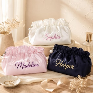 May include: Three satin pouches with gathered tops and pearl zipper pulls. One white pouch features the name Sophia in pink, one light pink pouch features the name Madeline in purple, and one navy blue pouch features the name Harper in gold.