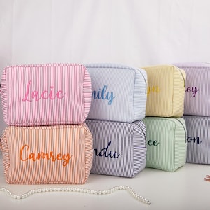 May include: A collection of striped cosmetic bags in various colors, including pink, orange, blue, yellow, green, and purple. Each bag features a different name embroidered in a complementary color. The bags are rectangular with a zipper closure.
