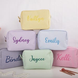 May include: A stack of six personalized cosmetic bags in various colors, including yellow, purple, blue, green, and pink. Each bag features a name embroidered in a complementary color, with a striped pattern.