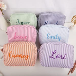 May include: Six personalized cosmetic bags in various colors, including green, pink, orange, blue, and purple. Each bag features vertical stripes and an embroidered name in a complementary color. The bags are designed for storing makeup and toiletries.
