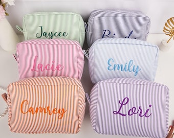 May include: Six personalized cosmetic bags in various colors, including green, pink, orange, blue, and purple. Each bag features vertical stripes and an embroidered name in a complementary color. The bags are designed for storing makeup and toiletries.