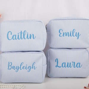 May include: Four light blue and white striped cosmetic bags stacked together. Each bag has a different name embroidered in blue: Caitlin, Emily, Bayleigh, and Laura. The bags have a rectangular shape with a zipper closure.