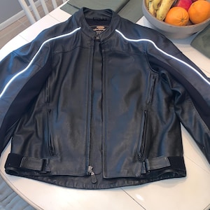 May include: Black leather motorcycle jacket with white reflective stripes on the sleeves and shoulders. The jacket has a zippered front, pockets, and a stand-up collar. A classic piece of outerwear.