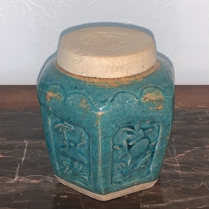 May include: A hexagonal ceramic jar with a teal glazed finish and a light beige lid. The jar features embossed floral patterns on each side, with visible wear and texture along the edges and rim, showcasing a rustic, weathered appearance.