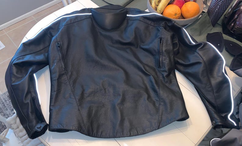 May include: Black leather motorcycle jacket with a mandarin collar and zippered pockets. Features white reflective stripes on the sleeves and shoulders. A classic design for motorcycle enthusiasts.