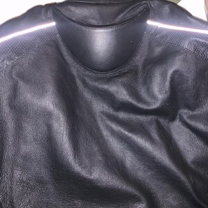 May include: Black leather jacket with a collar and reflective stripes on the shoulders. The jacket has a textured surface and a padded collar. The back of the jacket is visible, showing the stitching and design details.