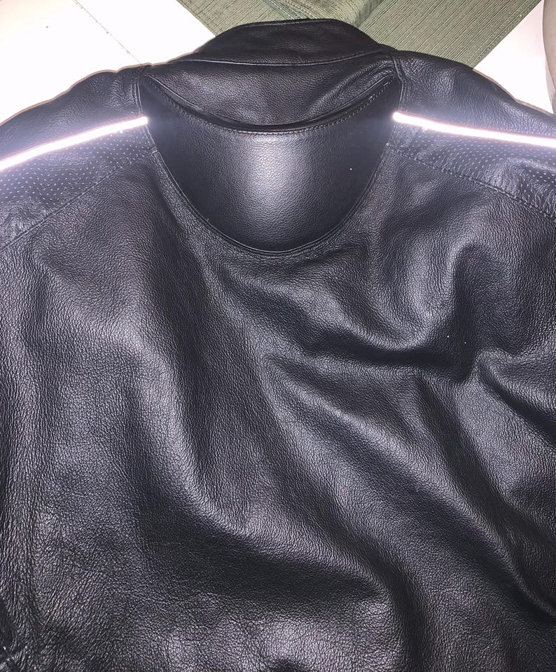 May include: Black leather jacket with a collar and reflective stripes on the shoulders. The jacket has a textured surface and a padded collar. The back of the jacket is visible, showing the stitching and design details.