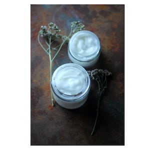 Op de afbeelding: Twee kleine, heldere glazen potjes gevuld met witte crème, tegen een rustieke, gevlekte bruine achtergrond. Gedroogde takjes van delicate, lichtgekleurde bloemen zijn rond de potjes gerangschikt en voegen een natuurlijk tintje toe. De crème ziet er glad en dik uit.