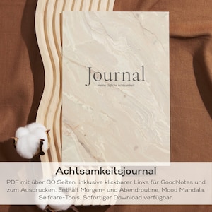 Könnte beinhalten: Ein Journal mit marmorähnlichem Einband, das Wort "Journal" ist in einer Serifenschrift gedruckt. Der Text "Meine tägliche Achtsamkeit" steht unter dem Wort "Journal". Das Journal liegt auf einer braunen Oberfläche. Der Text "Achtsamkeitsjournal" steht unten.