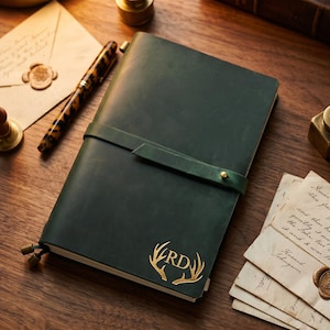 Op de afbeelding: Een donkergroen leren notitieboek met een bandsluiting en goudkleurige initialen "RD" met geweien. Een pen met een schildpadpatroon en briefpapier liggen in de buurt. Het notitieboek ligt op een houten oppervlak.