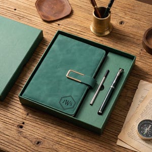 Op de afbeelding: Een groene journal met een gouden sluiting en de initialen "JWS" in een zeshoek, gepresenteerd in een bijpassende geschenkdoos met een pen en een navulling. De set wordt getoond op een houten oppervlak.