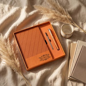Op de afbeelding: Een oranje geschenkset met een notitieboek, een pen en een penvulling, gepresenteerd in een bijpassende doos. Het notitieboek heeft een gestructureerde kaft met de naam "NICHOLAS" erop gedrukt. Een kleine keramische beker en verschillende beige notitieboeken zijn ook zichtbaar.