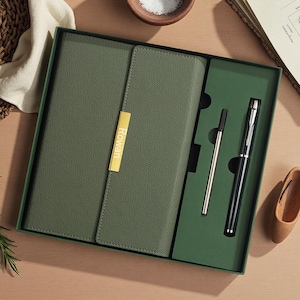Puede incluir: Un juego de diario y bolígrafo verde oliva en una caja de regalo. El diario tiene una placa dorada con el nombre "Rowan". El juego incluye un bolígrafo plateado y un bolígrafo negro. La caja es verde.