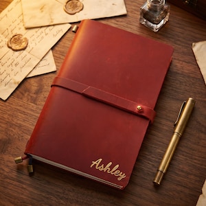 Op de afbeelding: Een rood leren notitieboek met een riemsluiting en de naam "Ashley" in gouden letters. Een gouden pen ligt naast het notitieboek op een houten oppervlak, samen met vintage brieven en een inktpot. Een klassiek schrijfwarenartikel.