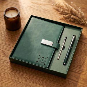 May include: A green leather journal and pen set in a gift box. The journal has a silver clasp and a debossed design with the initials "K S". A silver pen and a green pen are included. A candle sits to the left.