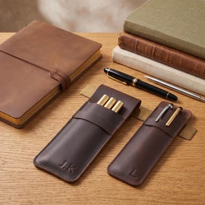 May include: A brown leather notebook, two brown leather pen cases with the initials J.K and L embossed on the front, three gold pens, a black fountain pen, and a silver pen. The pen cases are positioned on a brass ruler on a wooden surface.