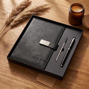 Könnte beinhalten: Ein schwarzes Lederjournal mit silbernem Verschluss und ein passendes Stift- und Kugelschreiber-Set, präsentiert in einer schwarzen Geschenkbox. Das Journal zeigt die Initialen "S.P" in einem Diamanten. Eine Kerze und dekoratives Gras sind im Hintergrund.