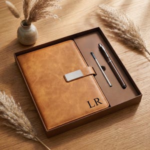 Op de afbeelding: Een bruin leren notitieboek met een zilveren sluiting en de initialen "LR" in zwart, gepresenteerd in een bruine geschenkdoos. De set bevat een zilveren pen en een bruine pen. Decoratieve droge planten en een kleine vaas staan op de achtergrond.