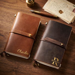Könnte beinhalten: Zwei Lederjournale, eines braun und eines dunkelbraun, mit goldenen Buchstaben und Riemen. Das braune Journal ist mit dem Namen "Charles" geprägt. Das dunkelbraune Journal ist mit dem Namen "Roman" geprägt. Beide Journale sind mit einem Lederriemen und einem Messingknopf verschlossen.