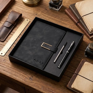 Op de afbeelding: Een zwarte leren notitieboekenset met een goudkleurige sluiting, een pen en een navulling in een zwarte doos. Het notitieboek is voorzien van de initialen "M.K." in een ruitvorm. Een leren penhouder en een liniaal zijn ook zichtbaar.