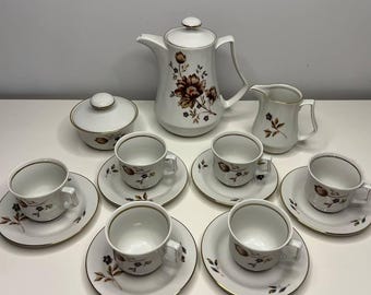 Könnte beinhalten: Ein weißes Porzellan-Teeservice mit floralem Design. Das Set beinhaltet eine Teekanne, eine Zuckerdose, einen Sahnekännchen sowie sechs Tassen und Untertassen. Das florale Muster zeigt braune und bordeauxfarbene Blumen mit goldenen Verzierungen.