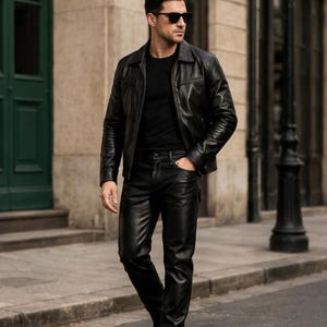 Op de afbeelding: Een zwart leren jasje en broek ensemble. De jas heeft een kraag en een knoopsluiting. De broek is getailleerd en heeft een klassiek ontwerp met vijf zakken. De outfit wordt gecompleteerd met zwarte schoenen en een zonnebril.