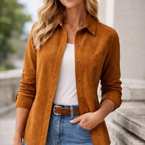 May include: A woman wearing a button-down, long-sleeved, brown suede shirt with two chest pockets, a brown leather belt, and blue jeans. The shirt is buttoned up and the sleeves are rolled up. She is also wearing a white tank top.