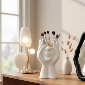 May include: A white ceramic makeup brush holder in the shape of hands and a face. The holder is filled with makeup brushes. A small white vase with dried flowers and a black-framed mirror are also visible.