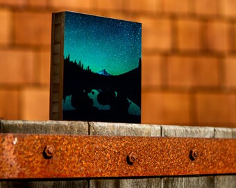 May include: A wooden block with a printed image of a mountain range under a starry night sky. The image is printed on a dark blue background with a green and blue gradient.