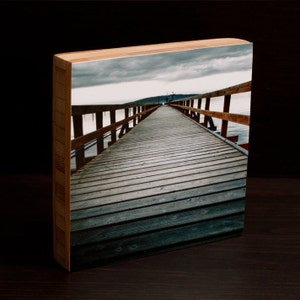 May include: A wooden photo block with a photograph of a wooden pier extending out into the water. The sky is overcast and the water is calm.