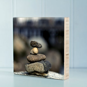 May include: A square wooden art piece featuring a stack of balanced stones. The stones vary in size, shape, and color, ranging from gray to brown. The background is blurred, suggesting a natural outdoor setting.