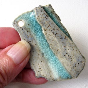 May include: A handmade ceramic tag with a hole, featuring a textured surface. The tag has a light blue stripe down the center and is speckled with dark brown spots. The edges are rough and irregular.