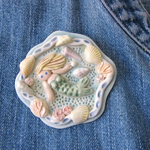May include: A handcrafted mermaid brooch with a light blue border, featuring a mermaid, seashells, and a seahorse. The mermaid has blonde hair and a green tail. The brooch is set against a blue denim background.