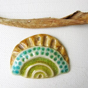 May include: A handmade ceramic pendant with a half-circle design. The pendant features a light brown, textured top section and a lower section with a green and turquoise pattern. A small hole is at the top for stringing. A dried leaf and stem are above the pendant.
