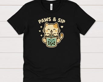 May include: Black t-shirt with a cartoon cat design. The cat, in light brown with tan spots, is drinking boba tea. The text "PAWS & SIP" is above the cat. The design includes hearts and stars, creating a cute and fun aesthetic.